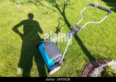 Ombre d'une personne debout près d'une tondeuse à gazon sur une pelouse verte pendant une journée ensoleillée, suggérant des soins de jardin et une activité de plein air. Banque D'Images