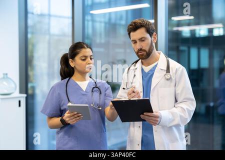 Deux professionnels de la santé examinent les dossiers d'un patient. L'un tient une tablette et l'autre utilise un presse-papiers. Ils semblent concentrés et engagés dans leur discussion. Banque D'Images