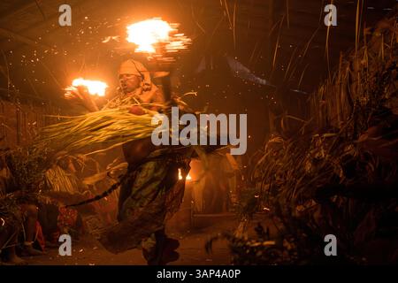 Les participants à une cérémonie des Bwiti, vêtus d'une tenue rituelle faite de feuilles de palmier séchées, dansent avec des torches allumées à l'intérieur d'un petit temple. Banque D'Images
