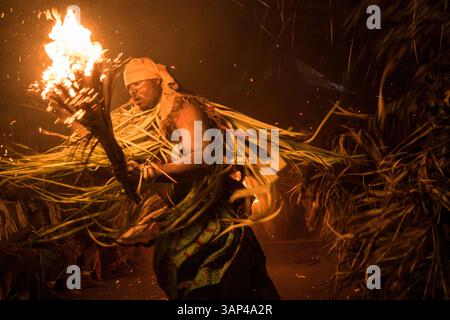 Les participants à une cérémonie des Bwiti, vêtus d'une tenue rituelle faite de feuilles de palmier séchées, dansent avec des torches allumées à l'intérieur d'un petit temple. Banque D'Images