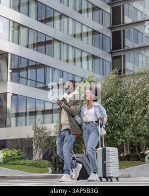 Couple afro-américain aimant, touristes avec des bagages marchant dans les rues de la ville se tenant la main et regardant l'environnement urbain. Banque D'Images