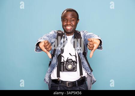 Homme afro-américain dans des vêtements décontractés, donnant joyeusement deux pouces vers le bas tout en se tenant sur fond isolé. Photographe masculin noir souriant avec appareil photo reflex numérique, exprime sa désapprobation en studio. Banque D'Images