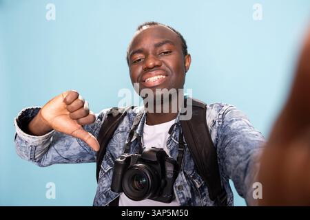 Heureux photographe afro-américain avec un appareil photo professionnel autour du cou, donnant les pouces vers le bas en studio. POV d'un vlogger masculin noir s'enregistrant tout en faisant un geste ludique de désaccord. Banque D'Images
