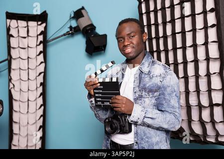 Cinéaste afro-américain debout dans le studio entouré de matériel d'éclairage, tenant une claquette. Portrait de photographe masculin noir avec appareil photo suspendu au-dessus de son cou et ardoise de film. Banque D'Images