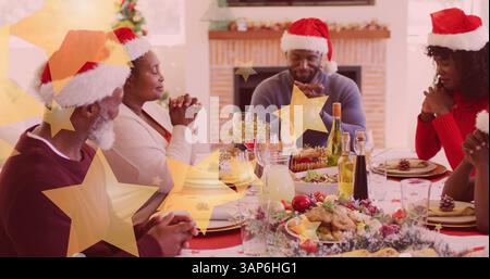 Image d'étoiles tombant sur la famille afro-américaine heureuse portant des chapeaux de père noël et priant Banque D'Images