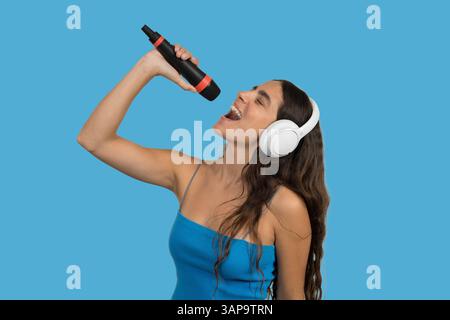 Chanteuse hispanique passionnée appréciant la musique, jouant avec un casque sans fil et un microphone en studio avec fond de couleur unie Banque D'Images