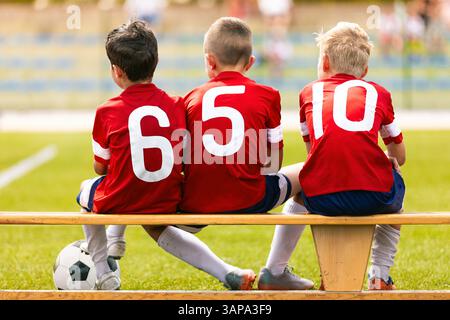 Les garçons en maillots de football rouges avec des numéros blancs sont assis sur un banc de substitution en bois, regardant le match, unis en équipe Banque D'Images
