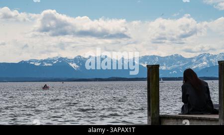 Starnberg, Allemagne. 16 avril 2025. Une femme est assise sur une jetée en bois sur le lac de Starnberg devant un panorama montagneux des Alpes bavaroises enneigées. Un petit bateau à moteur navigue sur le lac. Crédit : Leonie Asendorpf/dpa/Alamy Live News Banque D'Images