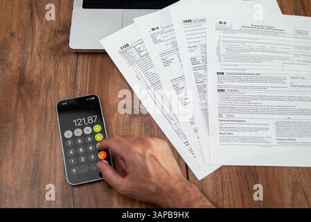 Homme entrepreneur compte le pourcentage d'impôts dans l'application mobile travaillant avec différents formulaires imprimés à la table Banque D'Images