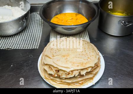 Des crêpes prêtes à l'emploi sont sur la table, dans une assiette blanche, à côté d'eux se trouvent des bols avec des produits pour faire des crêpes : jaunes, farine et beurre. Banque D'Images