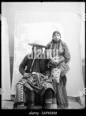 Cette image représente un homme et une femme amérindiens posés devant une couverture en toile de fond. L'homme est assis et vêtu d'une chemise euro-américaine, d'un chapeau de cow-boy et d'une couverture Pendleton, avec un étui de pistolet à sa ceinture. La femme se tient à côté de lui dans une robe d'aile géométrique avec châle Pendleton, boucles d'oreilles et accessoires perlés. La photographie fait partie de la collection Lee Moorhouse des bibliothèques de l'Université de l'Oregon. Banque D'Images