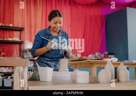 Une mexicaine, 24 ans, moule une pièce en céramique dans son atelier portant un tablier bleu, une chemise et un Jean. Des outils et des pièces finies l'entourent. Banque D'Images