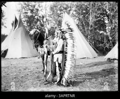 Un homme amérindien identifié comme 'Young Chief' se tient de profil, tourné vers la gauche, à côté d'un cheval de couleur foncée devant trois tipis. Le cheval est orné de perles géométriques, d'une selle et d'une bride perlée. L'homme porte un bonnet de traînée unique avec de l'hermine, une chemise en tissu, un gilet et des leggings. Il tient un tomahawk et un bandelier de fourrure. Le cheval et l'homme sont positionnés avec leurs ombres qui s'étendent derrière eux, face au soleil. Banque D'Images