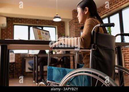 Employé féminin professionnel en fauteuil roulant examinant les rapports financiers sur ordinateur portable dans un lieu de travail moderne et inclusif. Femme asiatique handicapée travaillant sur la présentation de projet d'entreprise dans le bureau de démarrage. Banque D'Images