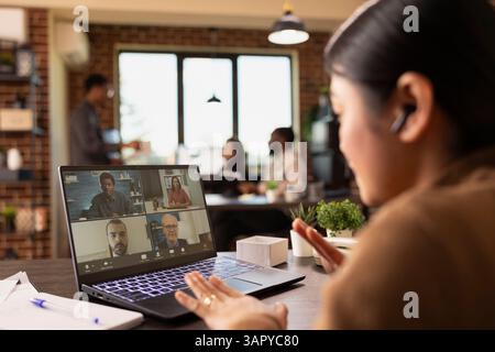 Gros plan d'un ordinateur portable affichant une vidéoconférence avec des collègues, tandis qu'une femme d'affaires avec des écouteurs s'engage dans la discussion. La manager asiatique assiste à une réunion en ligne depuis son bureau. Banque D'Images