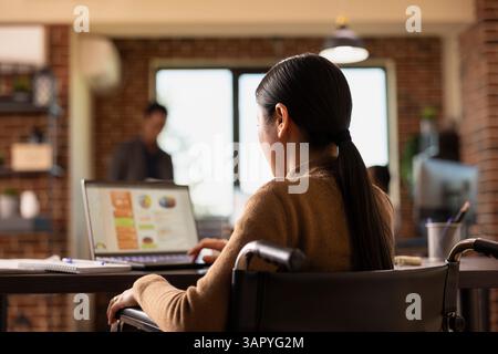 Femme d'affaires asiatique handicapée assise à son bureau avec son ordinateur portable et son ordinateur portable, analysant des graphiques financiers. Une employée féminine en fauteuil roulant rattrape le travail de recherche de l'entreprise dans le bureau de démarrage. Banque D'Images