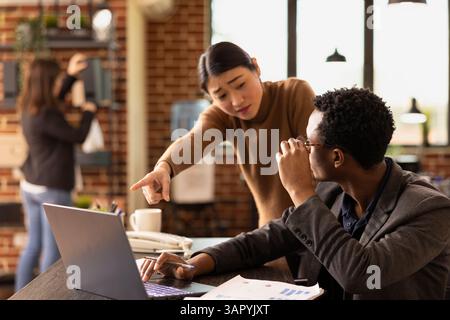 Entrepreneur masculin ajustant ses lunettes en tant que consultante féminine présente des statistiques financières sur appareil numérique. Les employés de start-up collaborent à la croissance de l'entreprise, examinent les rapports de l'entreprise sur ordinateur portable. Banque D'Images