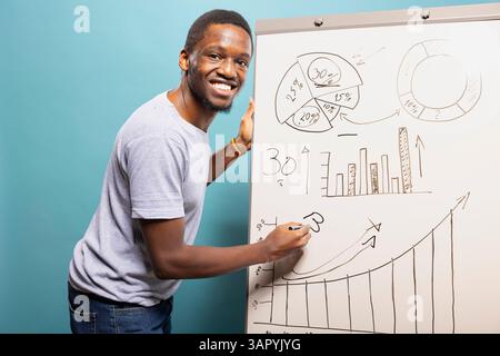 Jeune homme noir écrivant sur un tableau blanc et expliquant un graphique, debout sur un fond bleu de studio. Souriant afro-américain tenant un stylo et utilisant un tableau de marqueurs pour présenter un graphique. Banque D'Images
