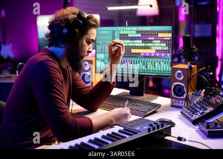 Compositeur auteur-compositeur prenant des notes pour créer une nouvelle chanson dans son home studio, écrivant les paroles et notes musicales avant d'enregistrer son morceau sur console de mixage et microphone, industrie de la musique. Banque D'Images