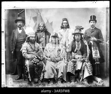 Photographie de Lee Moorhouse montrant sept hommes, dont deux identifiés comme John McBain et Lee Moorhouse, devant une toile de fond peinte avec des tipis. Les hommes sont habillés de costumes, et quelques personnages amérindiens sont présents. Banque D'Images