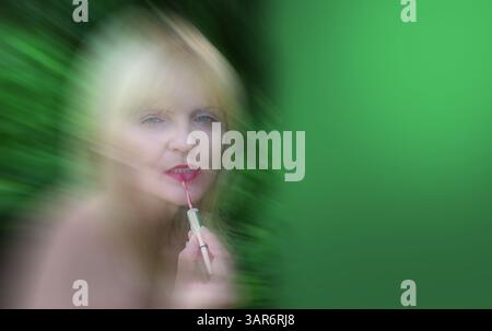 Fond vert avec une femme mature blonde floue appliquant de la crème de rouge à lèvres sur ses lèvres Banque D'Images