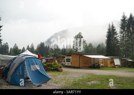 Camping avec une tente bleue et des cabanes en bois nichées dans un cadre de forêt brumeuse, entouré d'arbres verdoyants et de montagnes brumeuses, créant un Banque D'Images