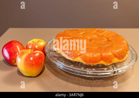 Tarte Tatin maison avec des tranches de pommes caramélisées dorées sur une assiette en verre clair, affichées à côté de pommes rouges et jaunes fraîches sur fond neutre Banque D'Images