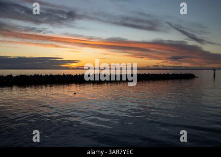 Un coucher de soleil serein se déroule sur des eaux tranquilles, projetant des reflets vibrants de teintes orange et violettes. Les roches silhouettées et les vagues douces améliorent le pe Banque D'Images