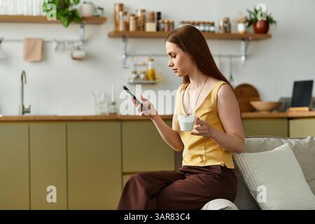 Une jeune belle femme se détend dans sa cuisine élégante, en sirotant un café et en utilisant son smartphone. Banque D'Images