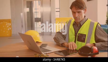 Image d'un ouvrier masculin caucasien utilisant un ordinateur portable et une tablette au bureau Banque D'Images