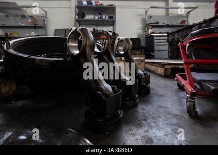 Les pistons avec bielles sont disposés sur le plancher d'un atelier de réparation ou d'un entrepôt, entourés par d'autres composants et outils de moteur. Banque D'Images