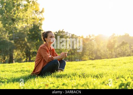 Femme détendue assise sur l'herbe dans le parc et tenant le téléphone portable. Banque D'Images
