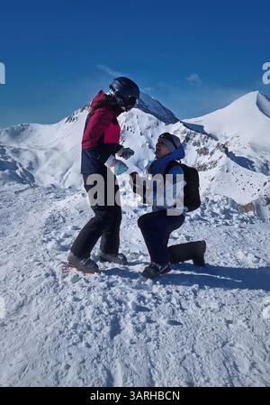 Proposition de mariage sur le sommet de la montagne enneigée comme un jeune homme sur son genou proposant le mariage avec une bague de mariage. Femme émotionnelle célébrant l'engagement. Banque D'Images