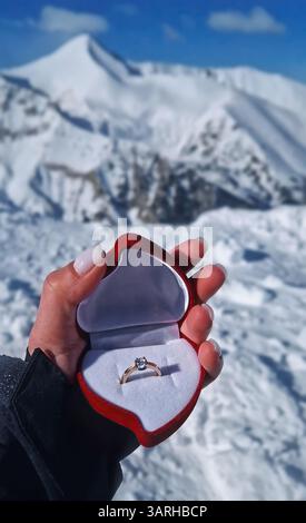 Main féminine gros plan tenant un anneau de fiançailles à l'intérieur d'une boîte en forme de cœur rouge sur le fond des sommets enneigés des montagnes. Proposition de mariage sur la monture Banque D'Images