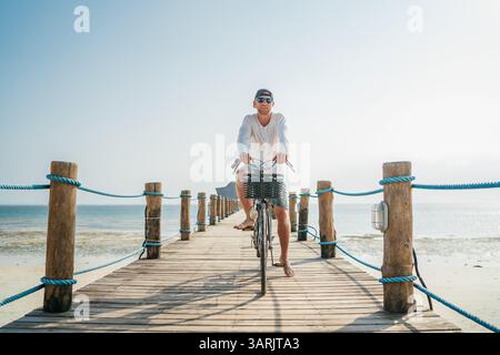 Portrait d'un homme heureux et souriant vêtu de vêtements d'été légers et de lunettes de soleil, à vélo sur une jetée en bois au bord de la mer. Des vacances sans soucis à Banque D'Images