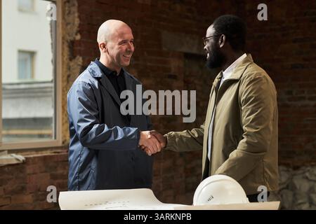 Plan moyen d'un contremaître caucasien et d'un architecte afro-américain se serrant la main tout en se saluant sur le chantier de construction Banque D'Images