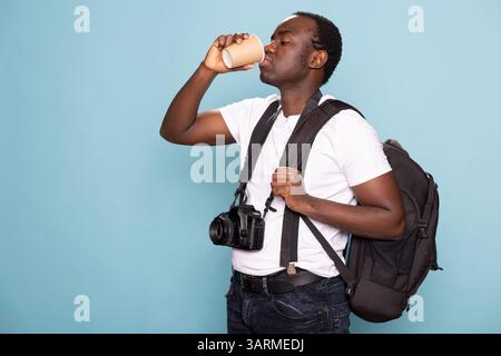Photographe tenant son sac sur son épaule et buvant du café dans un studio. Homme noir portant un t-shirt blanc avec bretelles, un appareil photo autour du cou, saisit son sac à dos et une tasse jetable. Banque D'Images