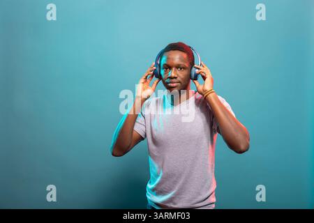 Portrait d'homme afro-américain portant des écouteurs sans fil et debout sur un fond isolé. Modèle masculin noir regardant la caméra en touchant son casque bluetooth, écoutant de la musique. Banque D'Images