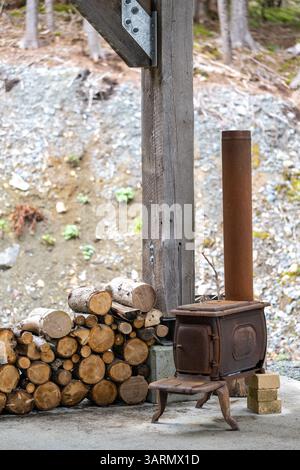 Un poêle à bois vintage avec une cheminée rouillée à l'extérieur près d'une aire de pique-nique. Les jonques en bois sont empilées à côté d'un poteau. Le poêle ventral du pot a de la rouille. Banque D'Images