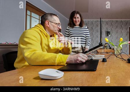 L'homme travaille sur des graphiques en utilisant un stylet à une table en bois avec un support pour ordinateur portable et une tablette numérique comme la femme souriante regarde. Banque D'Images