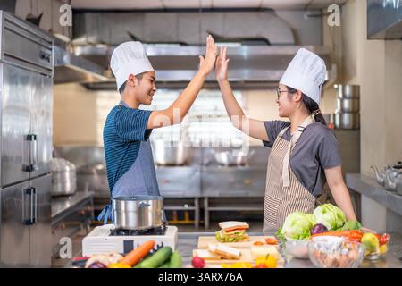 Deux jeunes chefs célèbrent avec un high-five dans une cuisine commerciale, portant des chapeaux de chef et des tabliers, montrant le travail d'équipe et la joie de cuisiner. Banque D'Images