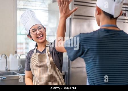 Deux jeunes chefs célèbrent avec un high-five dans une cuisine commerciale, portant des chapeaux de chef et des tabliers, montrant le travail d'équipe et la joie de cuisiner. Banque D'Images