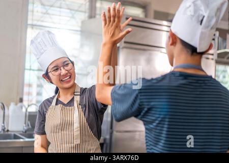 Deux jeunes chefs célèbrent avec un high-five dans une cuisine commerciale, portant des chapeaux de chef et des tabliers, montrant le travail d'équipe et la joie de cuisiner. Banque D'Images