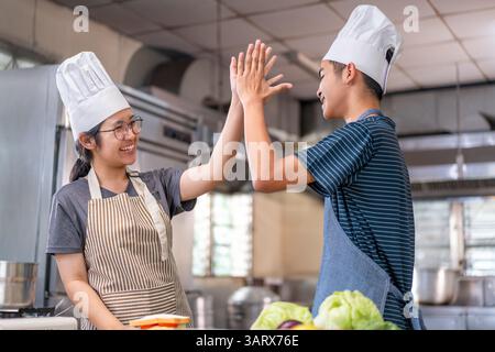 Deux jeunes chefs célèbrent avec un high-five dans une cuisine commerciale, portant des chapeaux de chef et des tabliers, montrant le travail d'équipe et la joie de cuisiner. Banque D'Images
