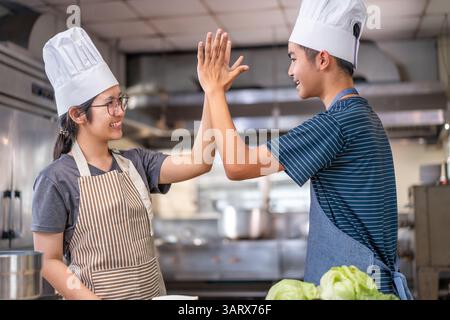 Deux jeunes chefs célèbrent avec un high-five dans une cuisine commerciale, portant des chapeaux de chef et des tabliers, montrant le travail d'équipe et la joie de cuisiner. Banque D'Images