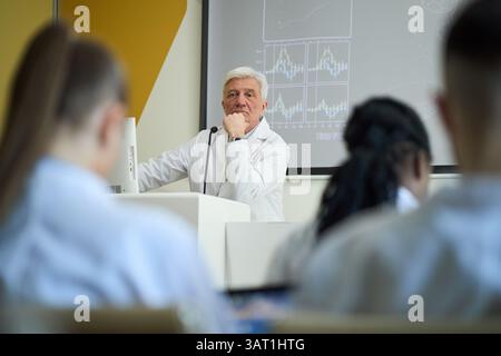 Scientifique principal présentant les résultats de la recherche avec divers graphiques et données à l'écran, divers étudiants écoutant attentivement dans la salle de conférence Banque D'Images