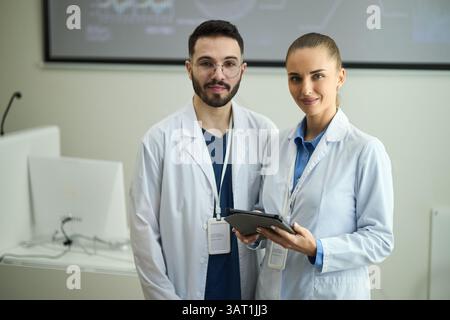 Portrait de deux professionnels de la santé souriants, un homme et une femme, portant des blouses de laboratoire blanches, debout dans un cadre clinique et tenant le comprimé devi Banque D'Images