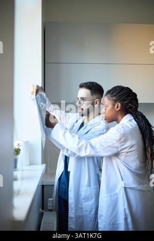 Deux professionnels de santé debout près de la fenêtre, examinant les radiographies du patient en milieu clinique Banque D'Images