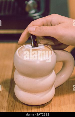 Gros plan d'une main remuant un cappuccino ou un latte dans une tasse blanche texturée unique sur une surface en bois Banque D'Images