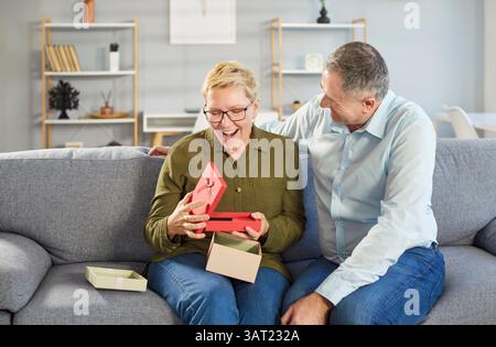 Heureux couple senior assis sur le canapé à la maison avec boîte cadeau cadeau célébrant les vacances. Banque D'Images